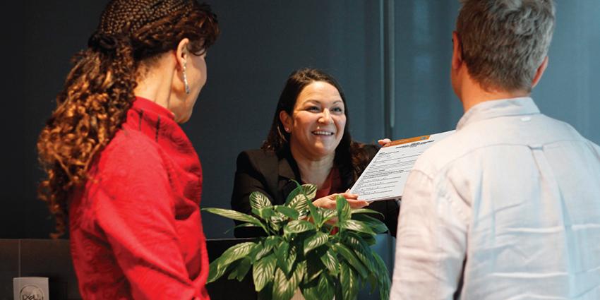 This shows a friendly interaction at what appears to be a customer service or reception desk. A woman, dressed professionally and smiling, hands over a document to a man and a woman standing on the other side of the counter. There is a plant on the desk, and the lighting is soft, creating a welcoming atmosphere.