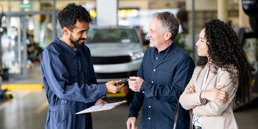 This image shows a positive interaction in an automotive service setting. A mechanic, dressed in a blue jumpsuit and holding a clipboard, is handing over car keys to a smiling male customer, who is accompanied by a woman.