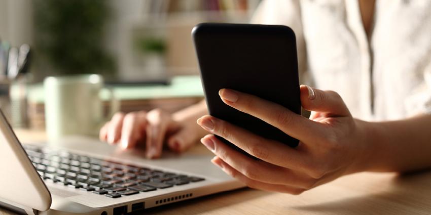 This close-up image captures a person using a smartphone while working on a laptop. The individual is seated at a desk with soft lighting, creating a focused and productive atmosphere.