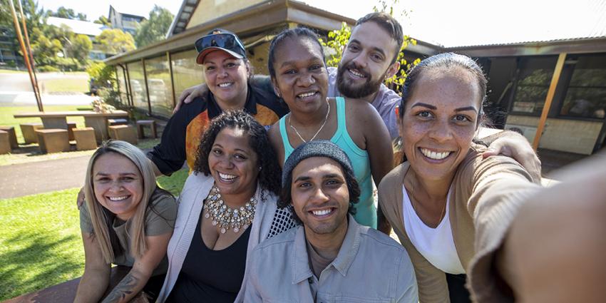 A group of people, smiling and taking a selfie outdoors. They appear relaxed and happy, gathered together in a casual setting with greenery and sunlight.